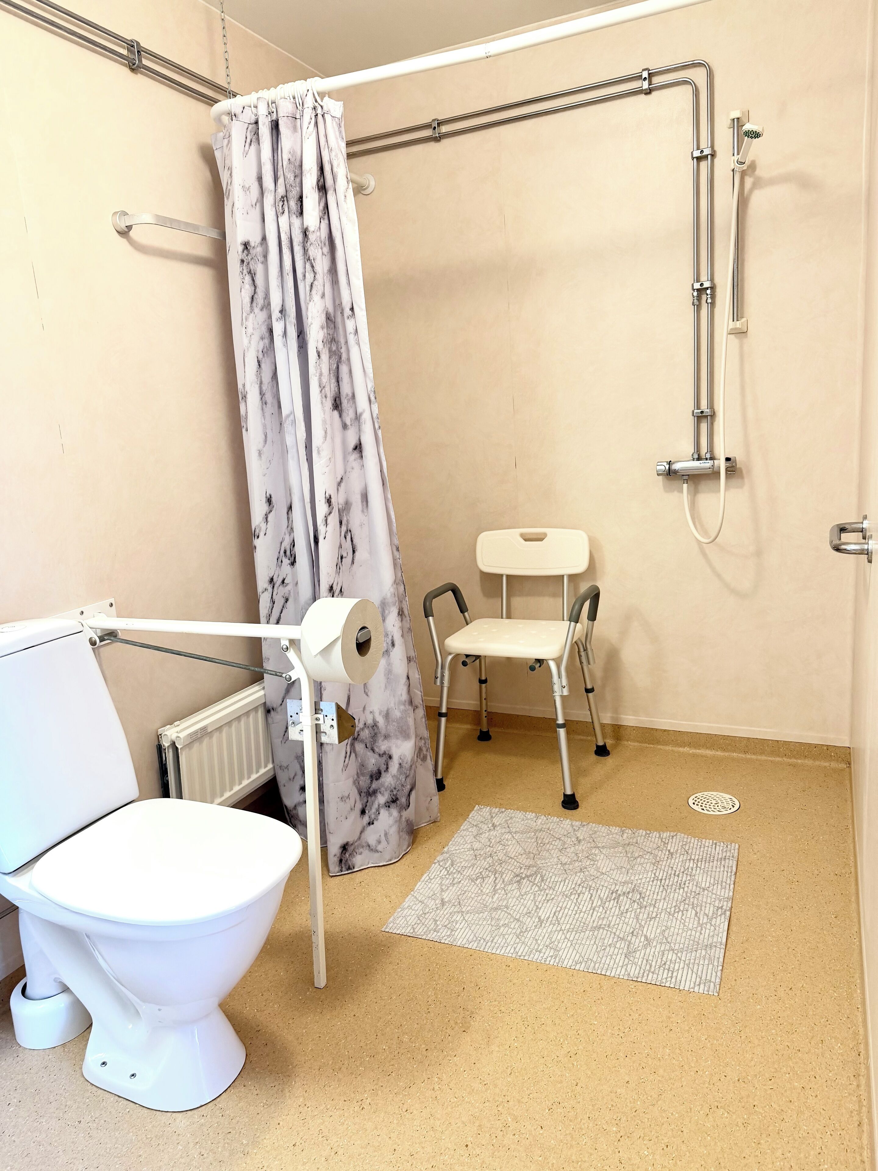 A bathroom with a toilet, a shower, a grab bar, a roll of toilet paper, a chair, and a mat.
