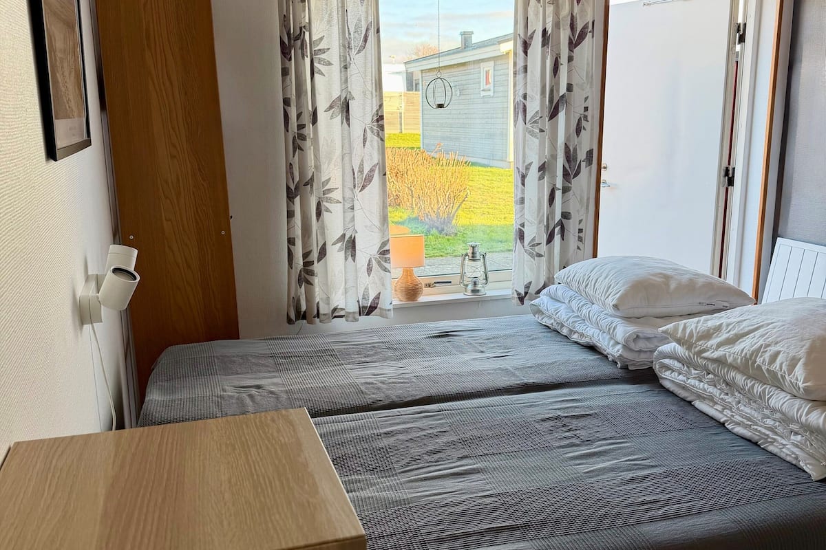 A bedroom with two beds, a wooden desk, a window with curtains, and a view of the outside.