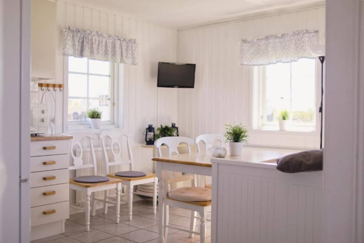 A bright, white room with a wooden table and chairs, a flat-screen TV mounted on the wall, and a window with a lace valance.