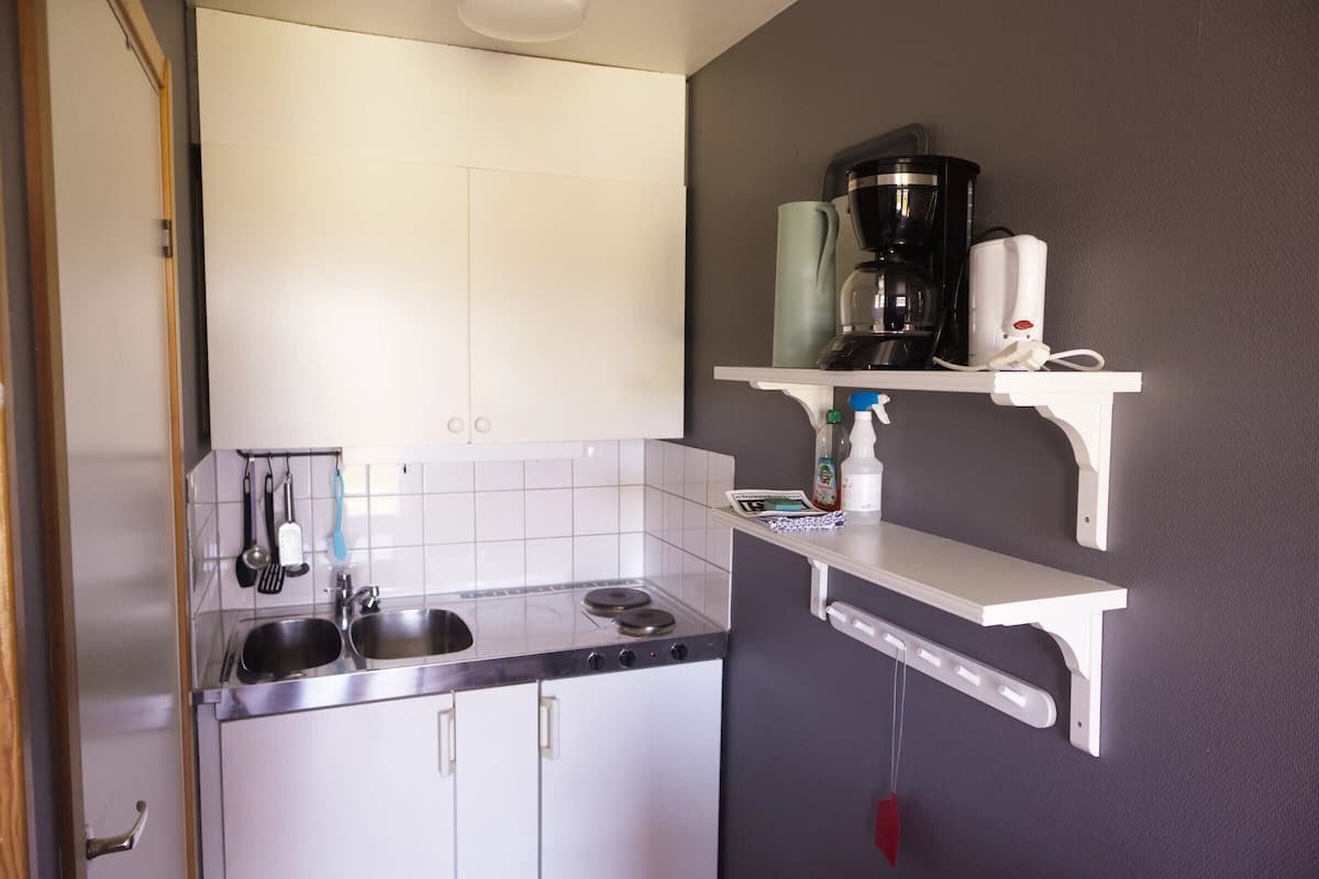 A kitchen with a stainless steel sink, two stovetops, white cabinets, and a white countertop. There are hanging utensils and a coffee maker on a shelf.