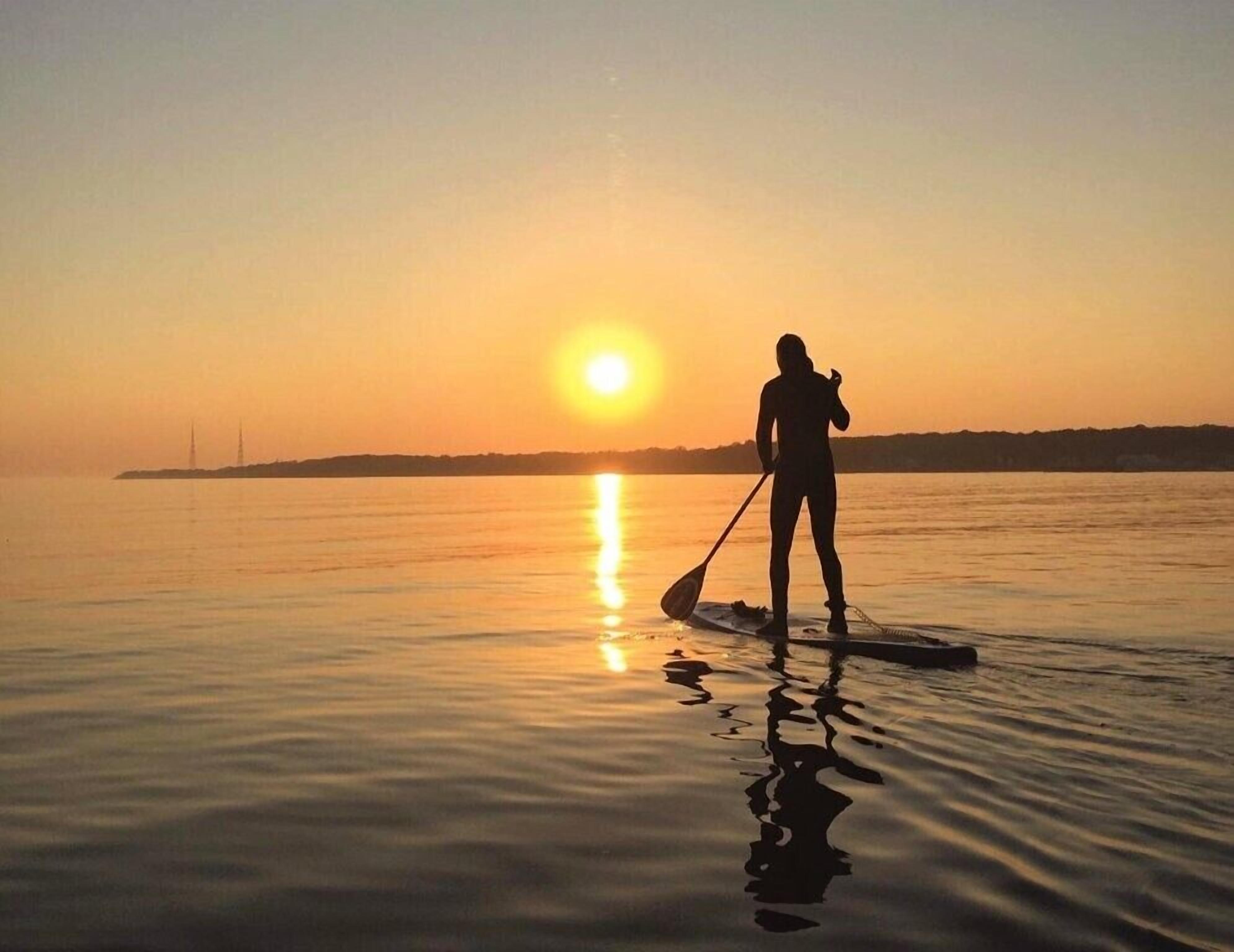 Una persona haciendo paddleboard al atardecer, con agua tranquila y una orilla lejana.