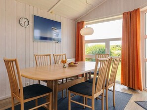 A dining area with a wooden table, four chairs, a clock, a painting, and a window with curtains. - 4 Star Holiday Home in Friedrichskoog (Friedrichskoog)