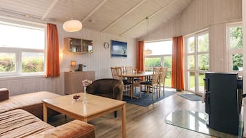 Amplia sala de estar con sofá, mesa de centro, mesa de comedor con sillas, chimenea y ventanas grandes con cortinas.