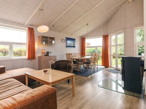 A spacious living area with a sofa, a coffee table, a dining table with chairs, a fireplace, and large windows with curtains. - 4 Star Holiday Home in Friedrichkoog (Friedrichskoog)