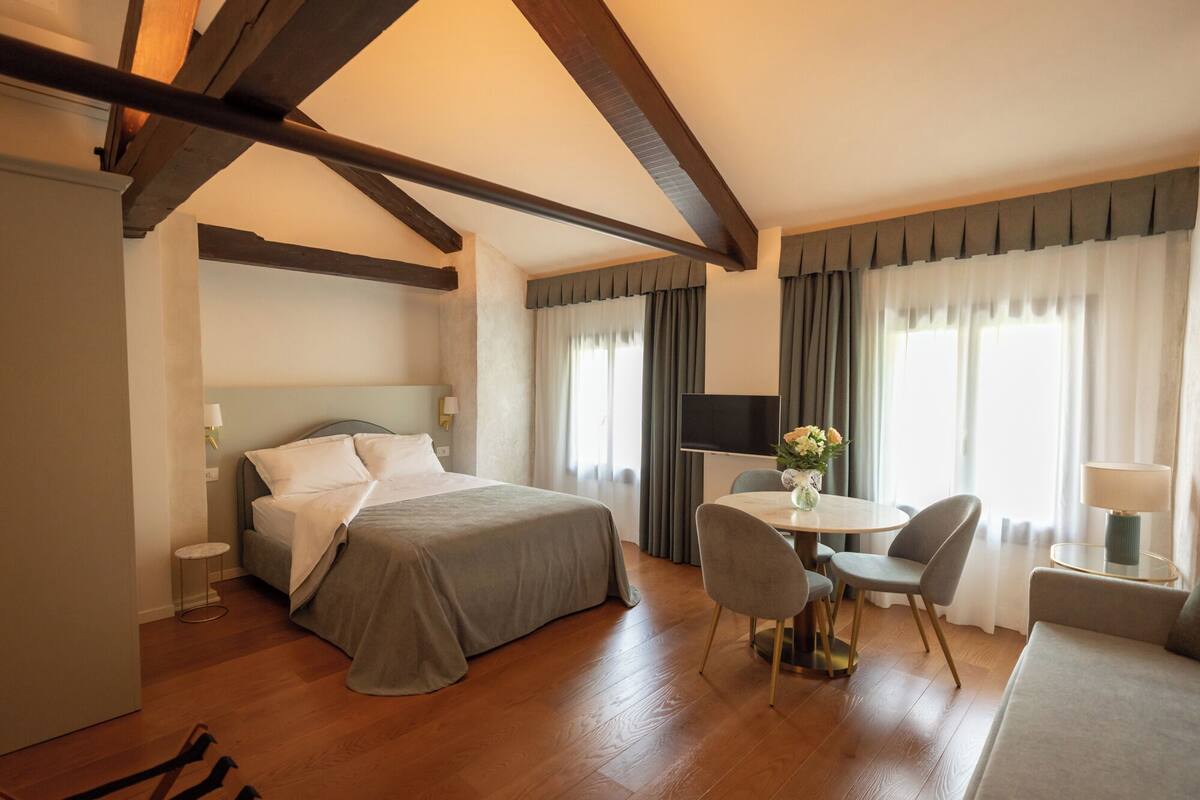 A hotel room with a bed, a small round table with chairs, a TV, and a window with curtains.
