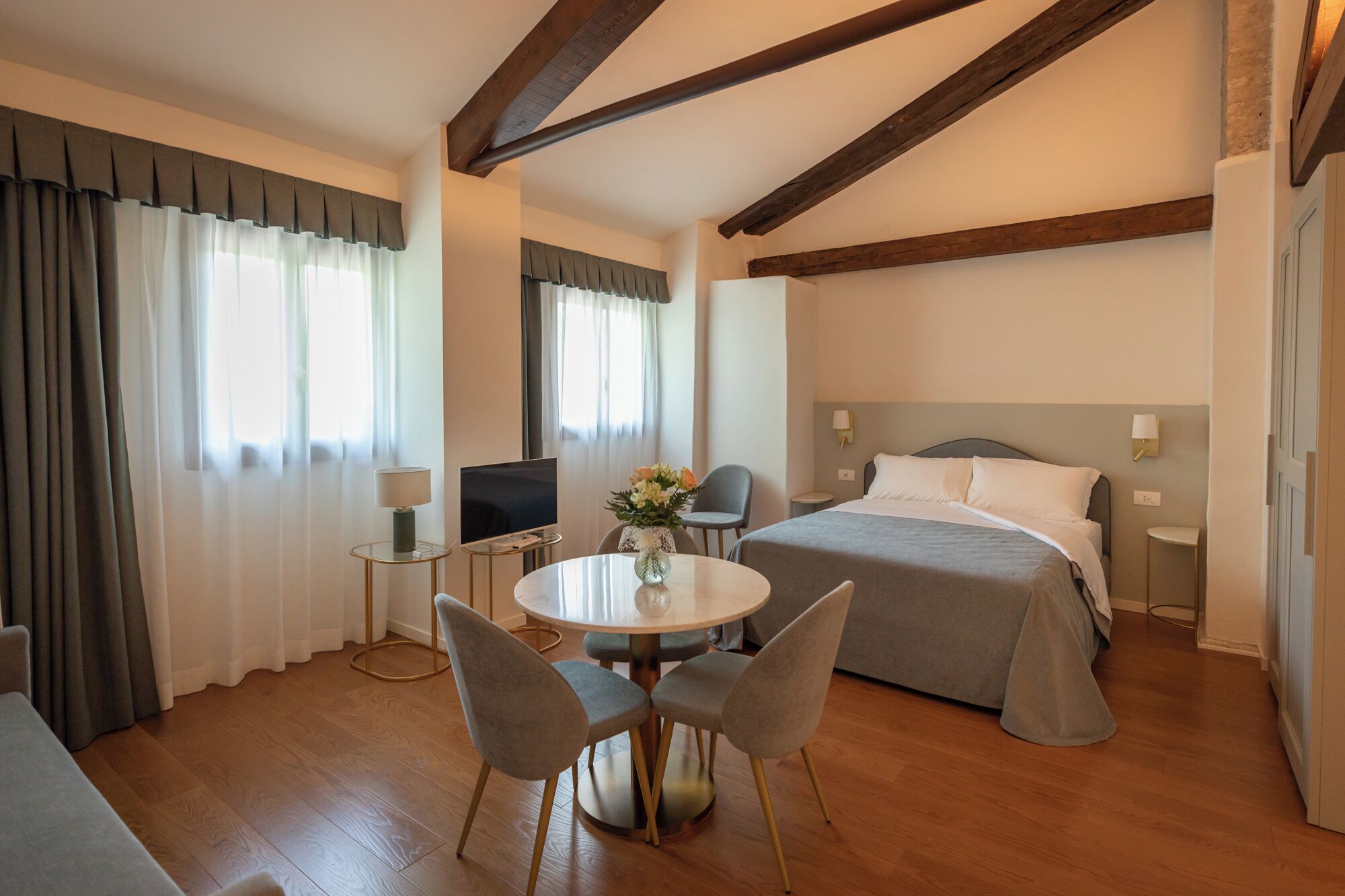 A hotel room with a bed, a TV, a desk with a lamp, and a dining area with chairs.