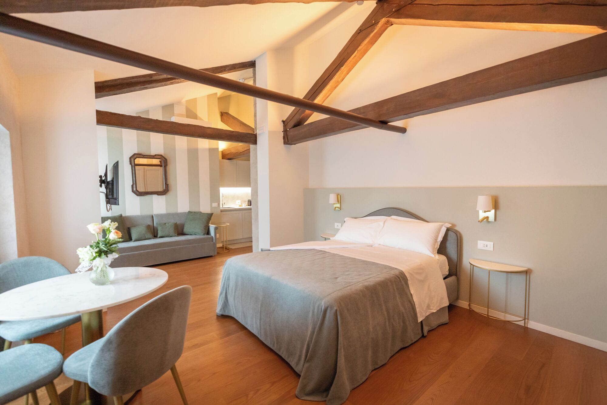 A bedroom with a bed, a small table, and a sofa. There is a kitchen in the background.