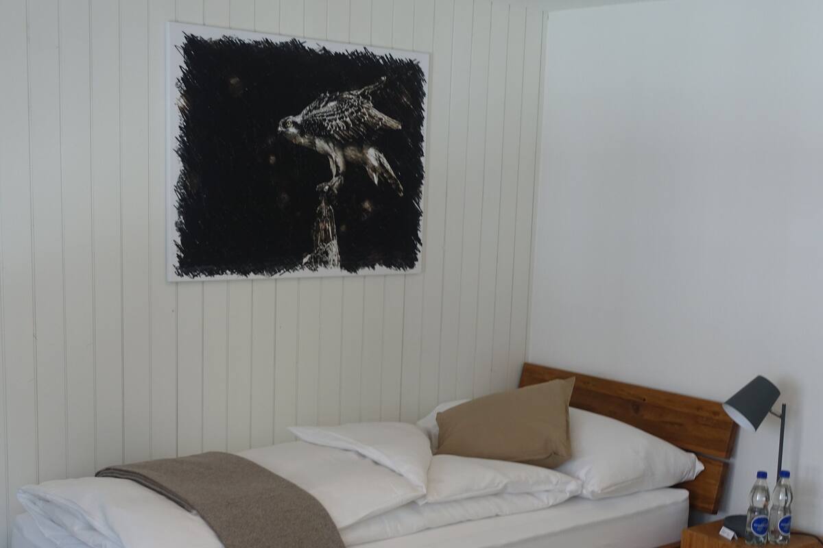 Una cama bien hecha con estructura de madera, cabecera y mesita de noche con lámpara y dos botellas.