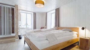 A modern bedroom with a wooden bed, white linens, a ceiling light fixture, and large windows. - Hotel Bären Guttannen (Guttannen)