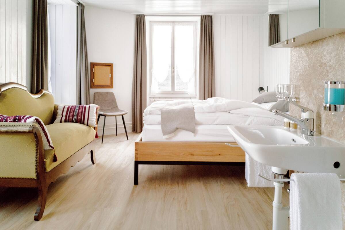 Una habitación de hotel con cama, una silla, una mesita de noche, un ventanal grande y un baño con lavamanos y espejo.