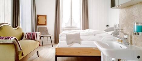A hotel room with a bed, a sofa, a chair, a large window, a sink, and a mirror.
