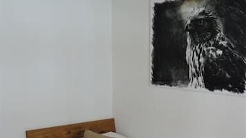 Un lit simple avec une tête de lit en bois, une literie blanche et un plaid gris. Une bouteille d’eau est posée sur une table de chevet en bois, à côté du lit. Un tableau abstrait en noir et blanc est accroché au mur, au-dessus du lit.