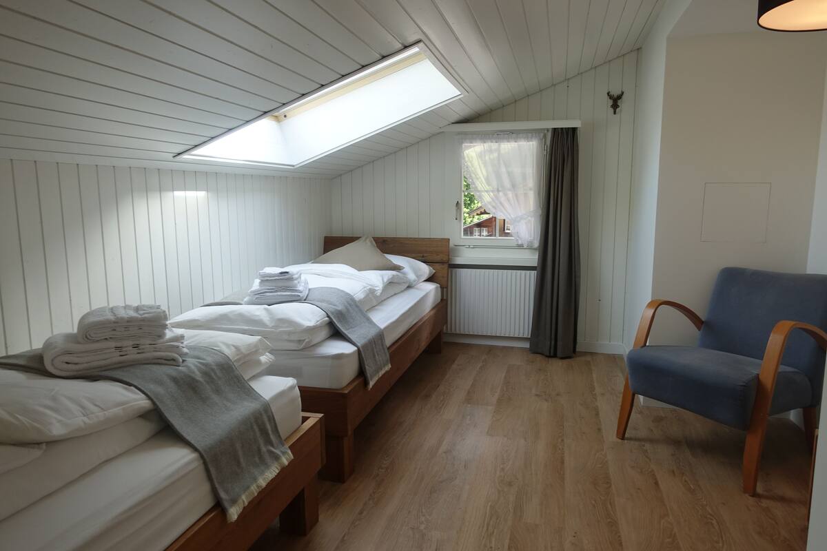 Una habitación con dos camas, una ventana con cortinas, una silla y un tragaluz.
