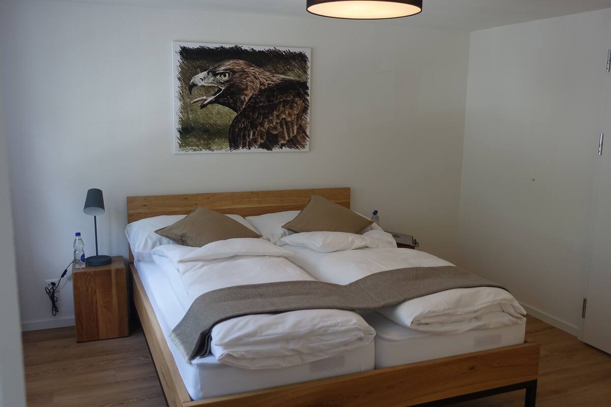Una cama bien hecha con ropa blanca y un cabecero de madera, una mesita de noche con una lámpara, y un cuadro de un pájaro en la pared.