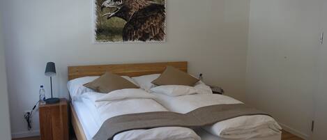 A neatly made bed with white linens and a wooden headboard, a bedside table with a lamp, and a painting of a bird on the wall.