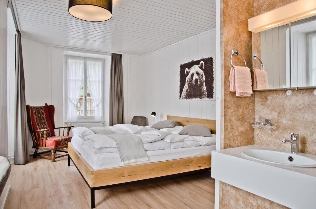 Un dormitorio con cama de madera, una silla, un ventanal con cortinas, un espejo montado en la pared y un baño con lavabo y toallero.
