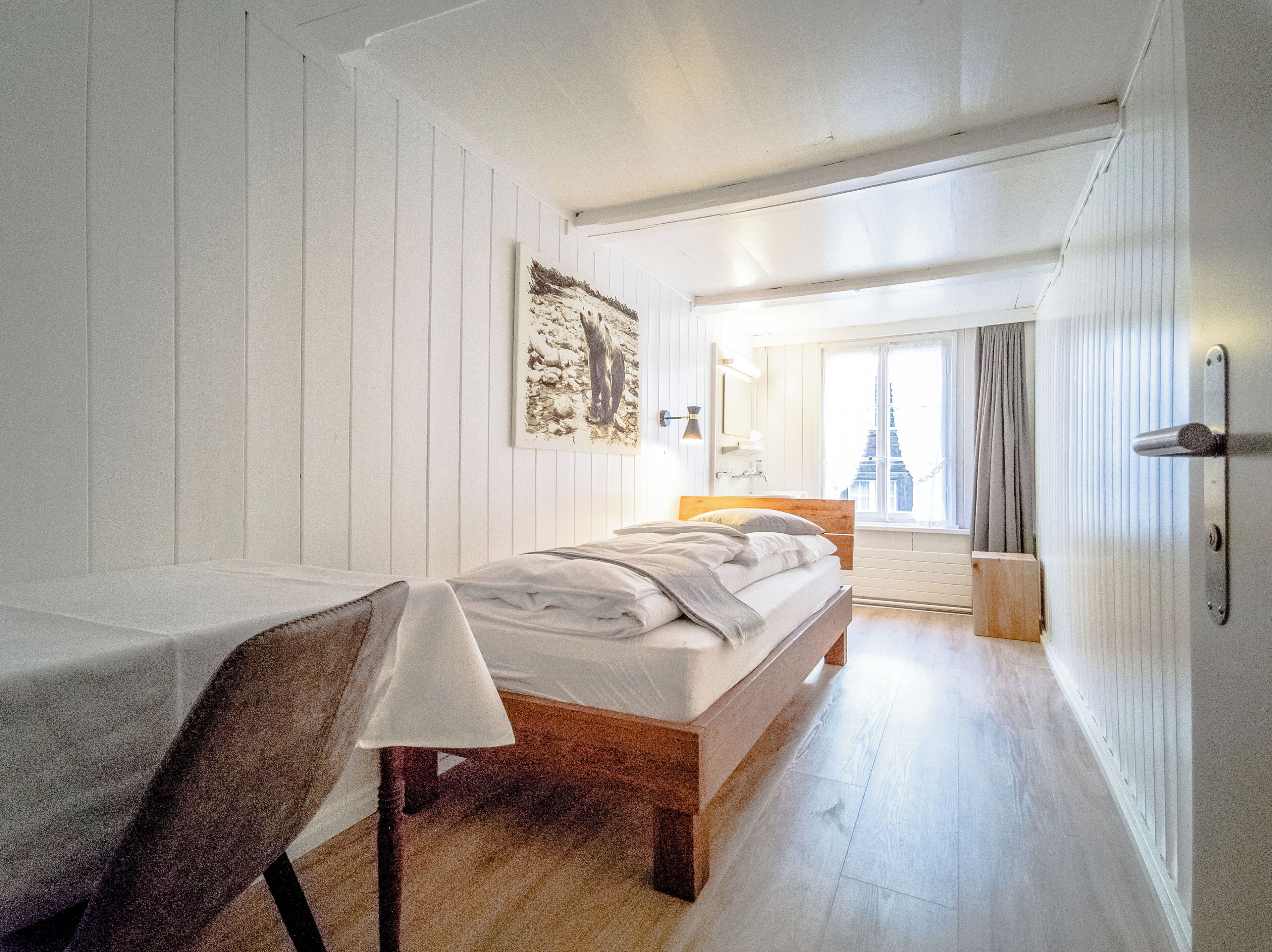 A bedroom with a bed, a chair, a bedside table, a wall-mounted lamp, and a window with curtains.