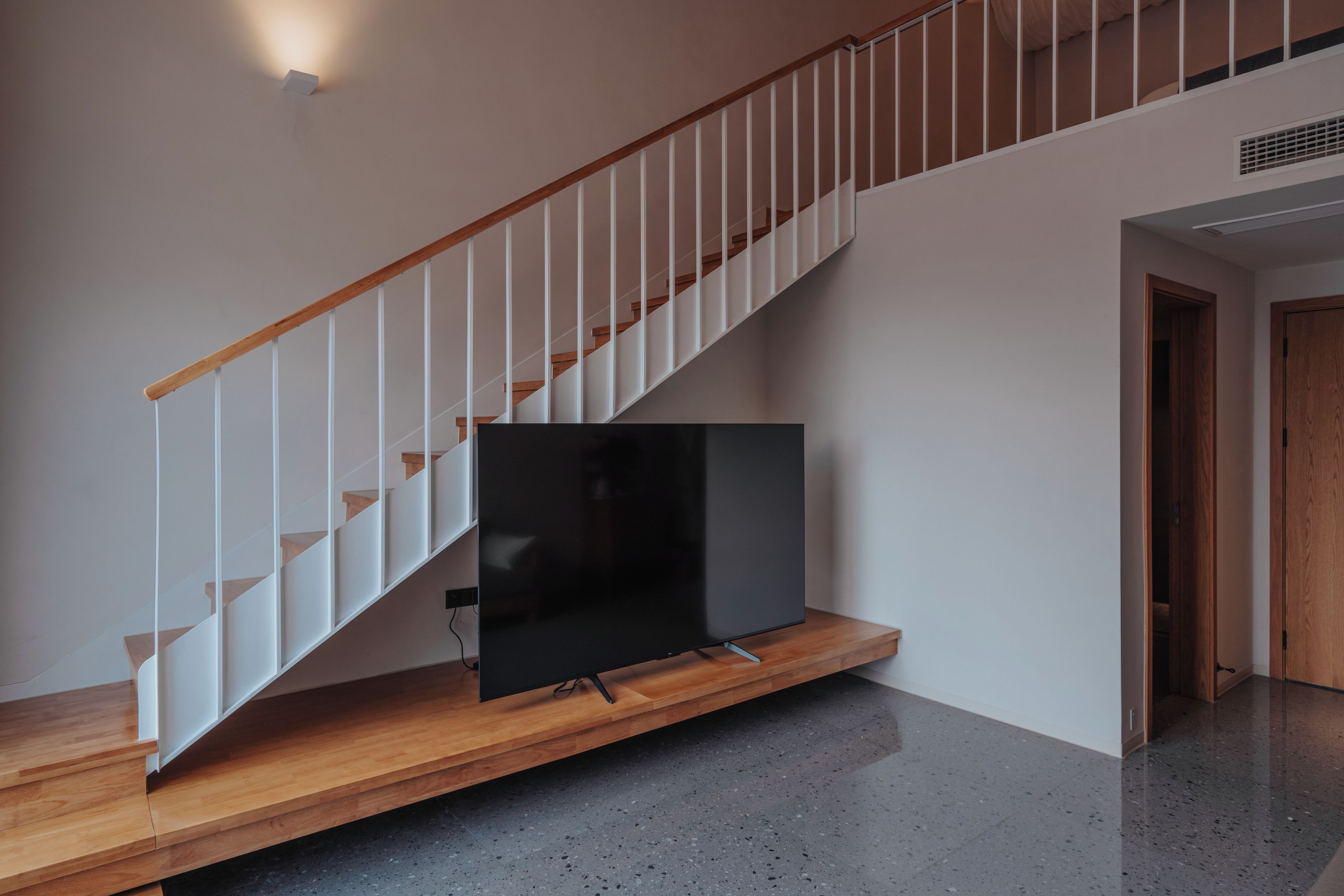 A modern staircase with wooden steps and white railings, leading to a landing with a flat-screen TV mounted on a wooden platform.