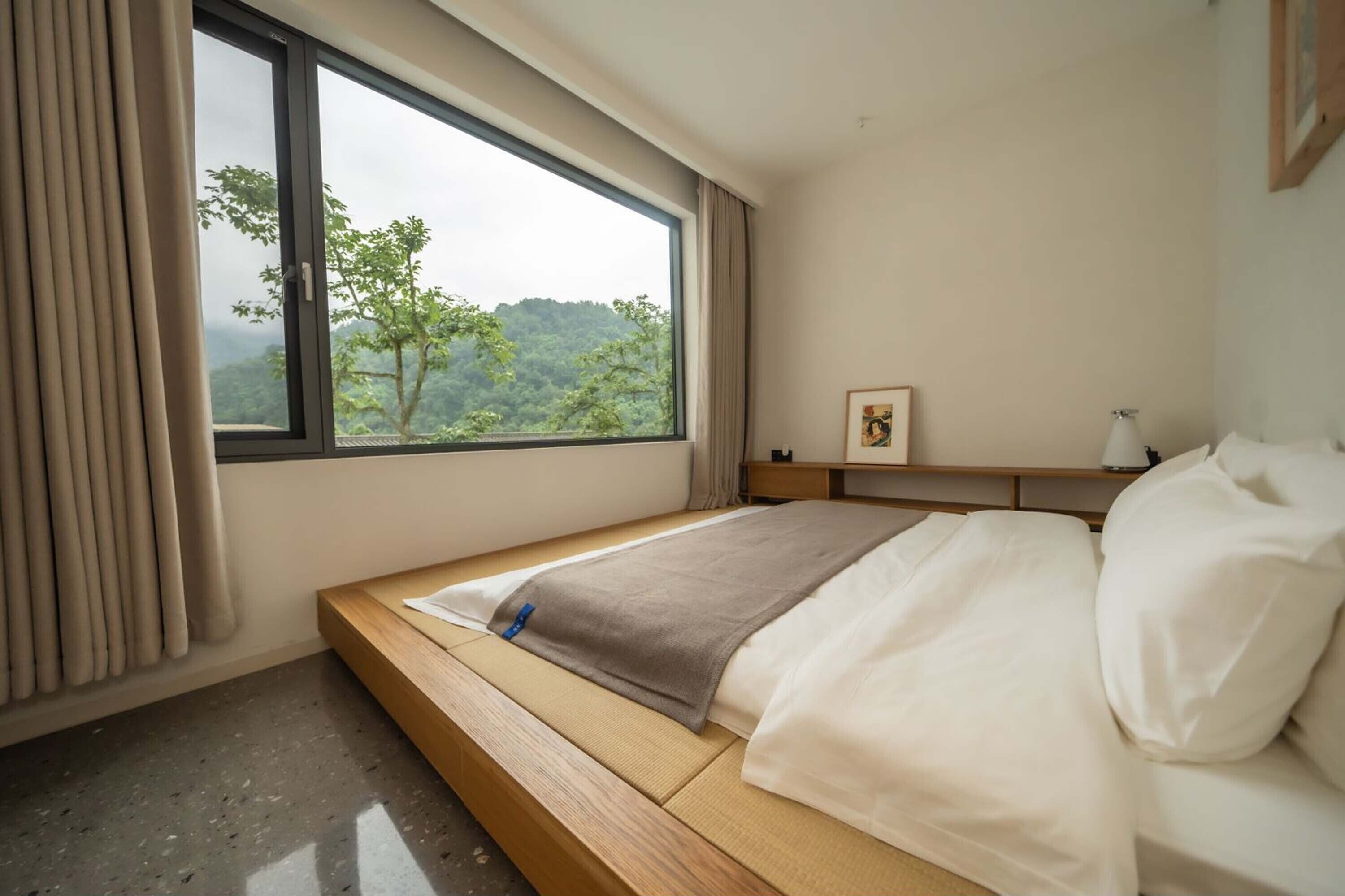 A bedroom with a bed, a window with a view of greenery, a bedside table, and a nightstand.