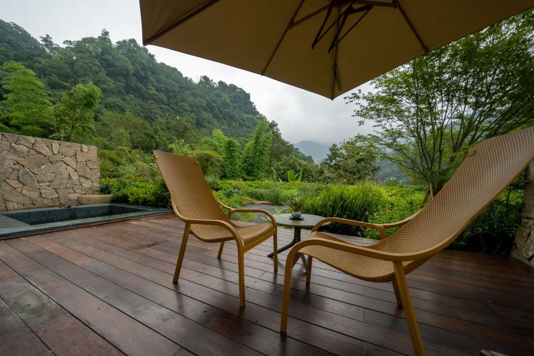 An outdoor patio with wicker chairs and a table, surrounded by a stone wall and lush greenery.