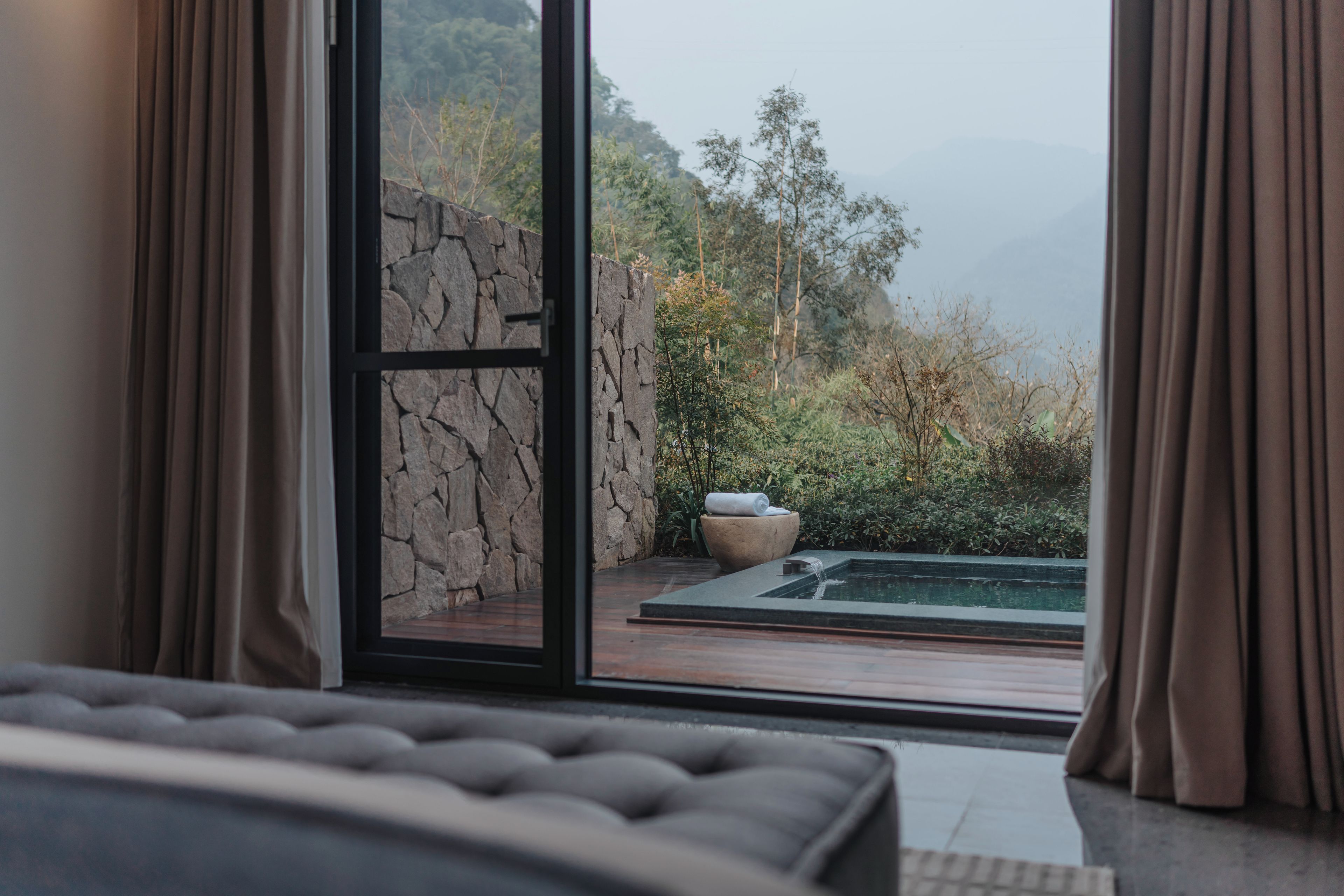 A bedroom with a large window, a stone wall, and a view of a garden with a water feature.