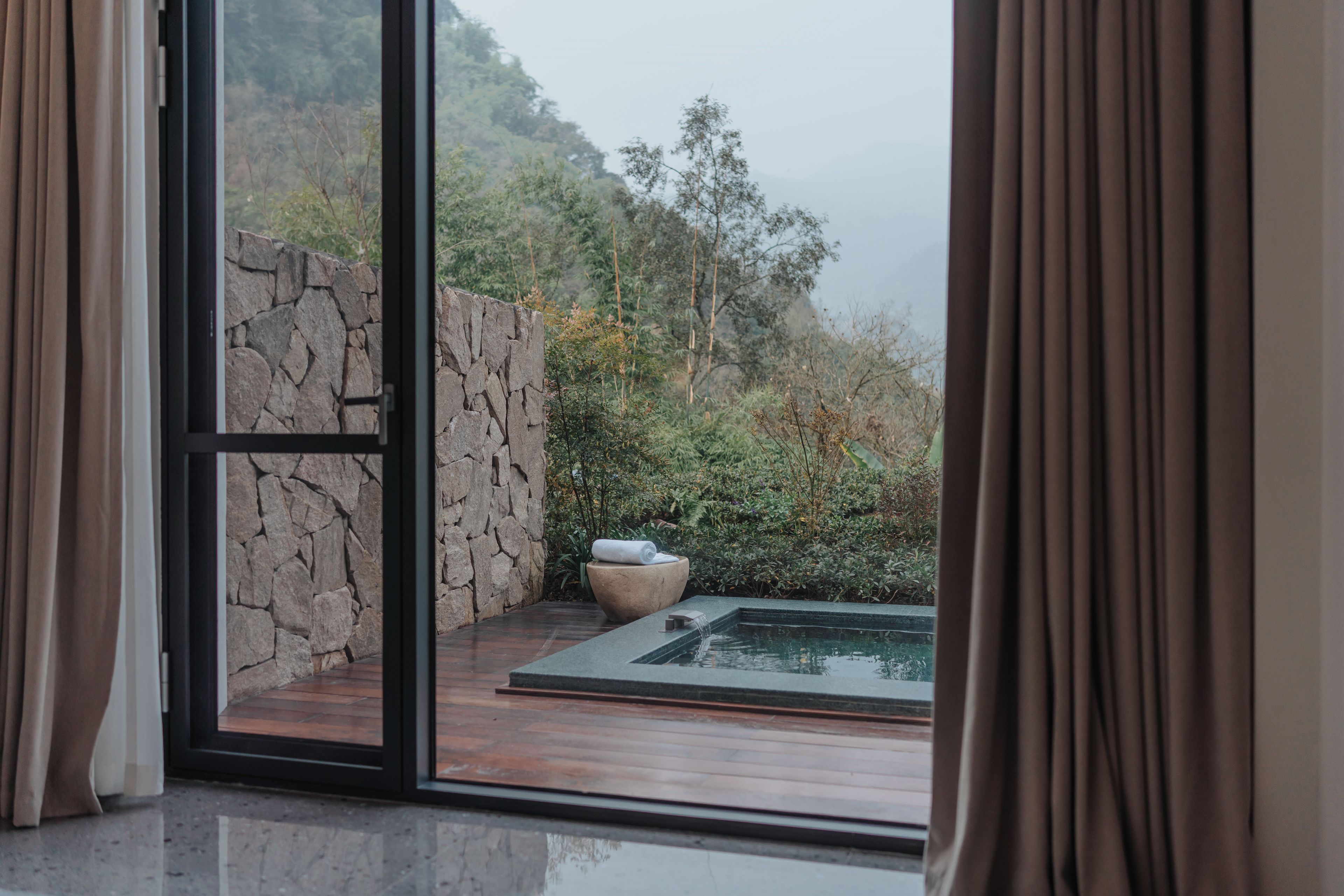 A room with a stone wall, a wooden floor, and a view of a garden with a water feature.