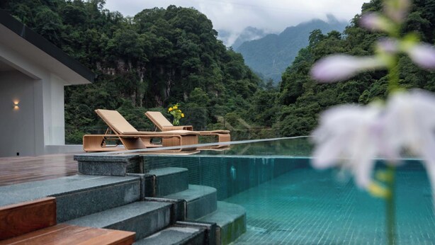 Un área de piscina moderna al aire libre con escalones que conducen a sillas de descanso, rodeada de abundante vegetación y montañas.