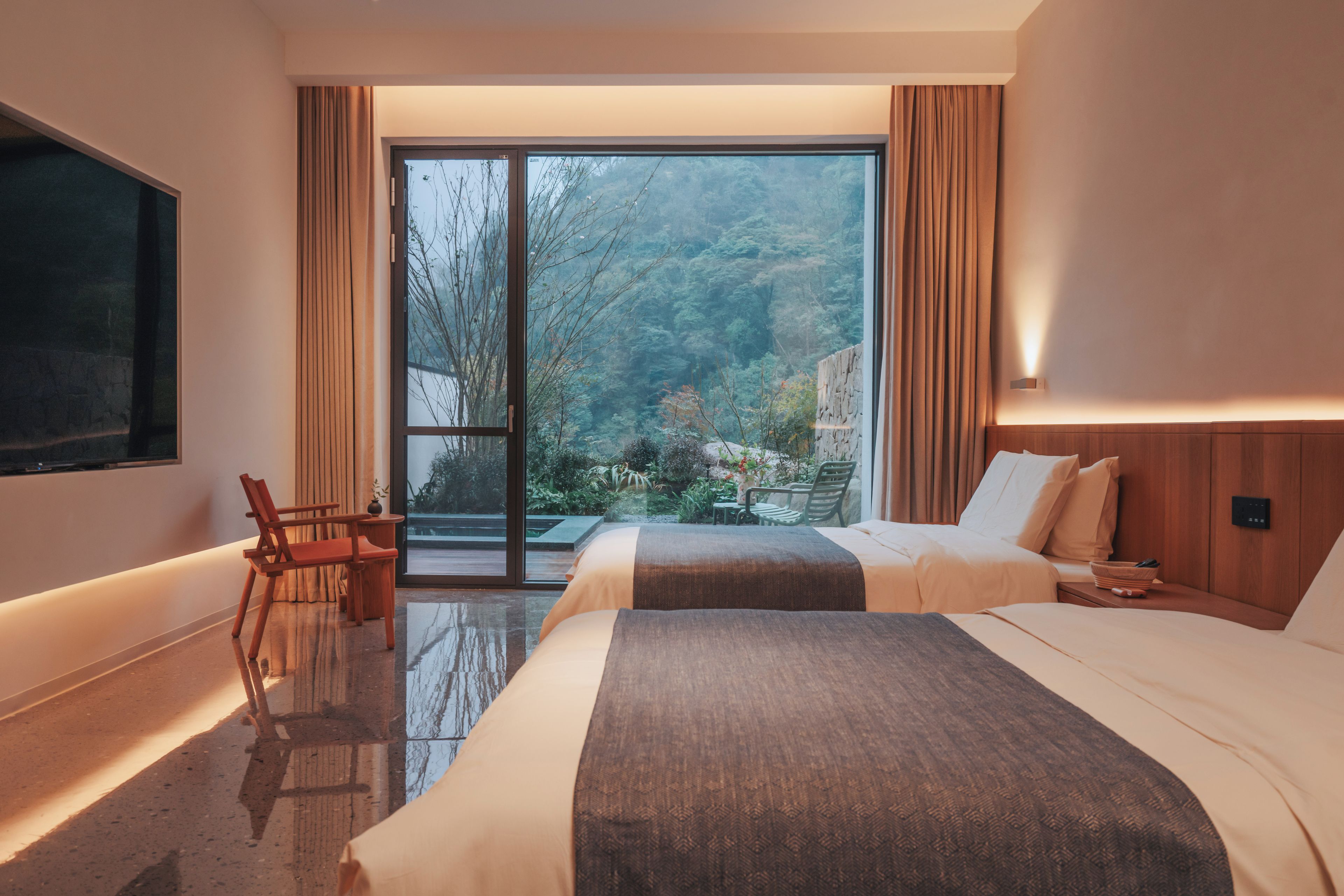 A hotel room with two beds, a desk, a chair, and a large window offering a view of a forested area.