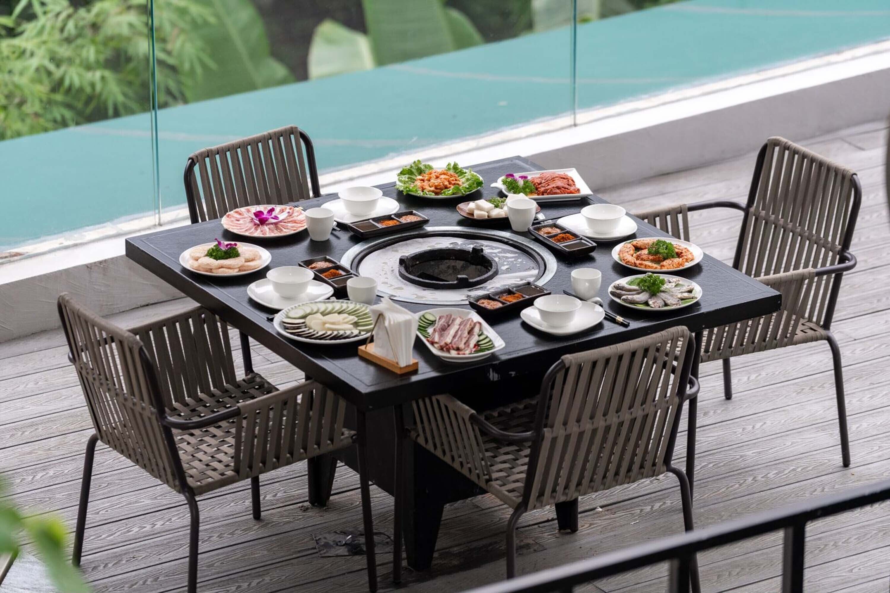A dining table set with various dishes and a central grill, surrounded by wicker chairs on a wooden deck with a glass railing and a view of greenery.
