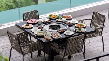 A dining table set with various dishes and a central grill, surrounded by wicker chairs on a wooden deck with a glass railing and a view of greenery.