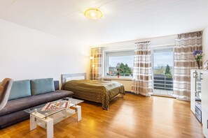 Una sala moderna con un sofá, una mesa de centro de vidrio, una cama con cubrecama estampado y vistas al exterior a través de una ventana.