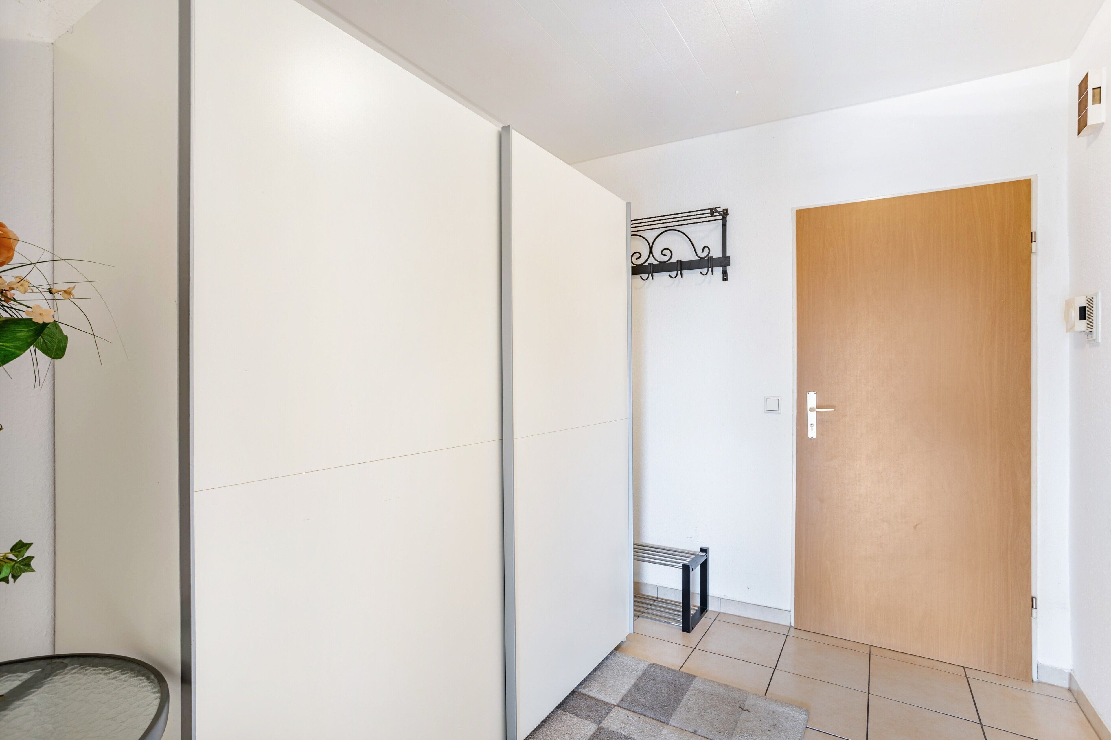 Un pasillo con puerta de madera, decoración metálica en la pared, un pequeño banco y una mesa de vidrio.
