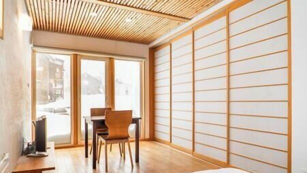 Une chambre moderne équipée d’un lit, d’un bureau et d’une chaise. On y trouve une porte coulissante à motifs en bois et une fenêtre donnant sur la rue.