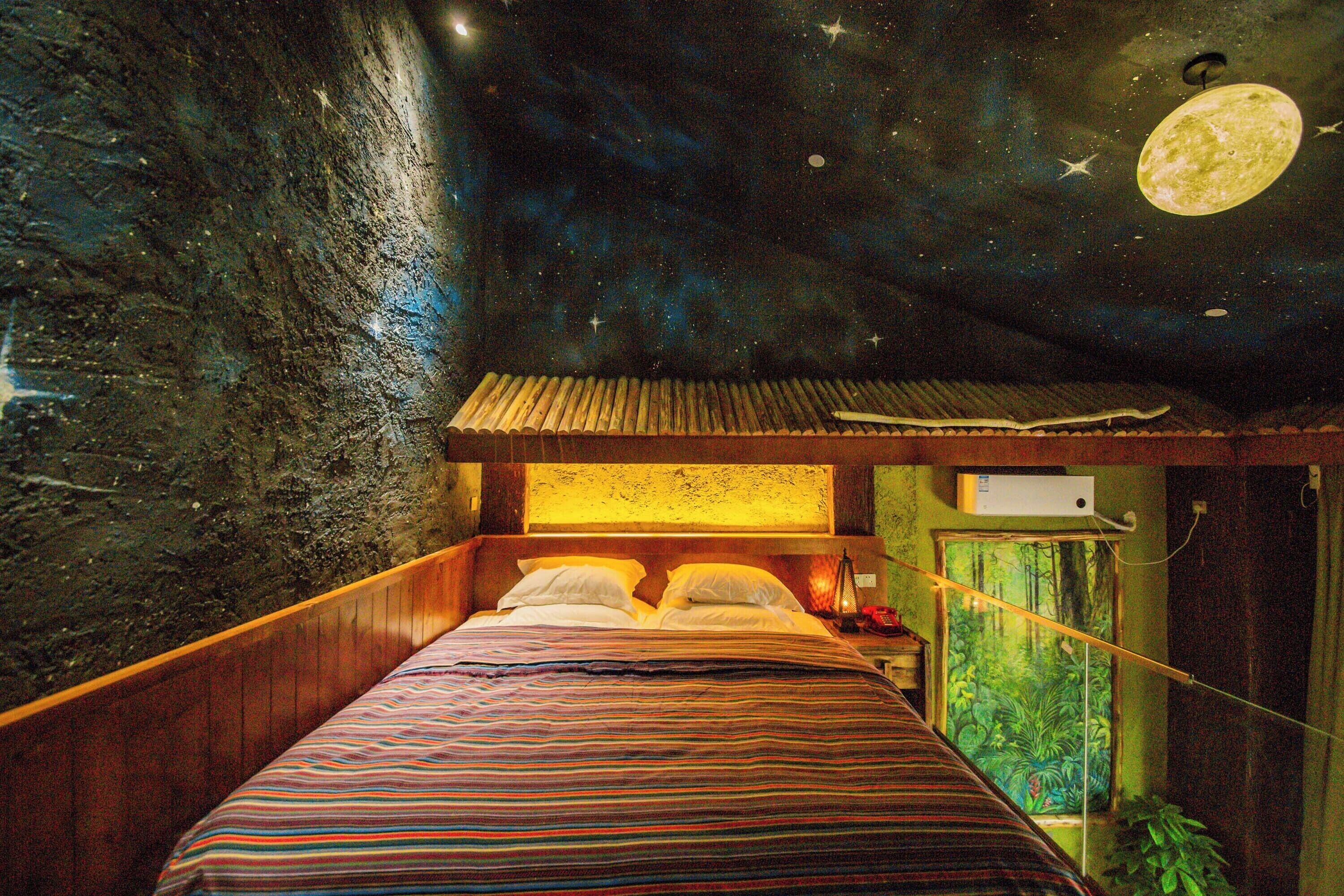 Un lit recouvert d’une couvre-lit à rayures, une fresque représentant un ciel étoilé au plafond, et une lune figurant dans cette fresque.