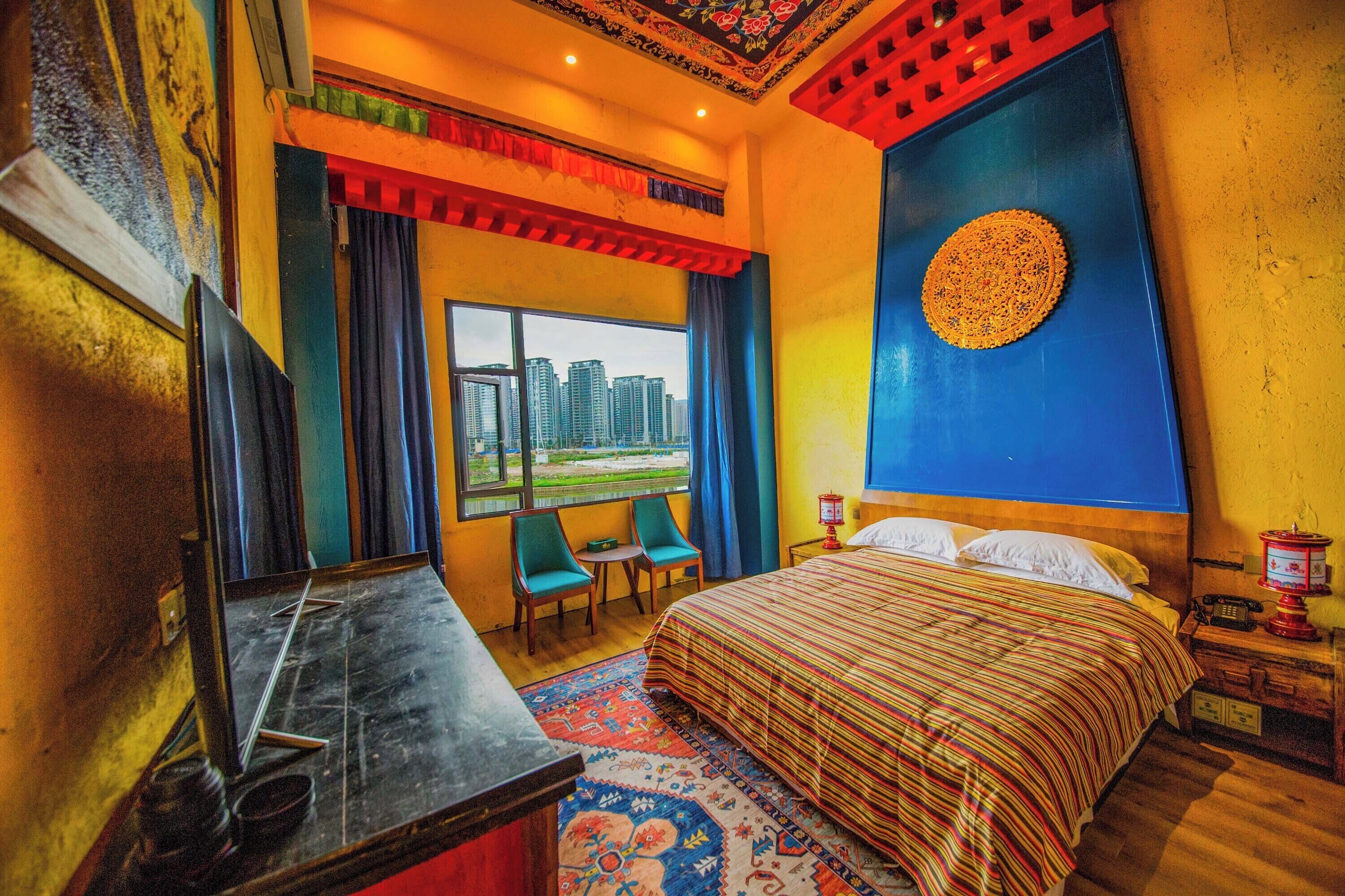 A hotel room with a large bed, a desk with a mirror, a small table, two chairs, a window with a city view, and a decorative wall piece.
