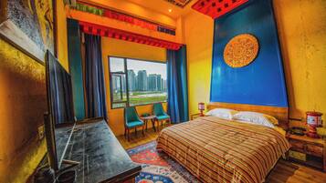 A hotel room with a large bed, a desk with a mirror, a small table, two chairs, a window with a city view, and a decorative wall piece.
