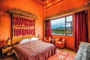 Une pièce comprenant un lit, deux chaises, une table, une fenêtre donnant sur un plan d’eau et des bâtiments, et un mur décoré de piments rouges.