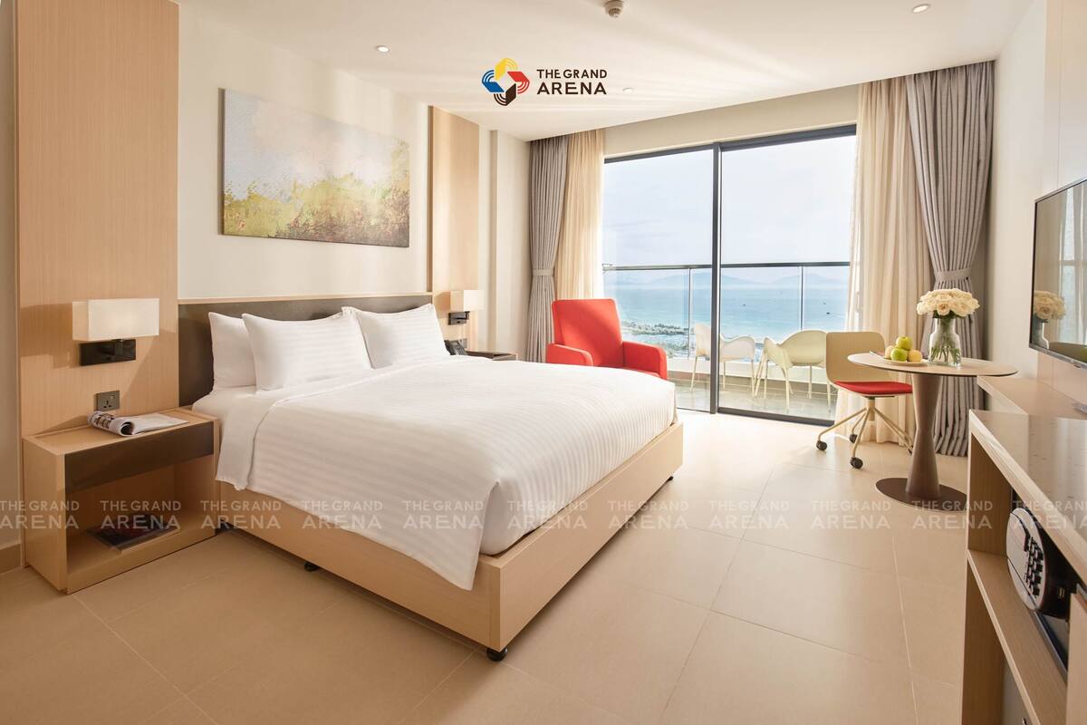 A hotel room with a large bed, a desk with a chair, a TV, and a view of the sea.