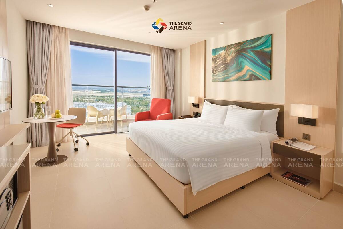 A hotel room with a large bed, a desk, a chair, a small table with flowers, a red chair, a painting, and a view of the city.