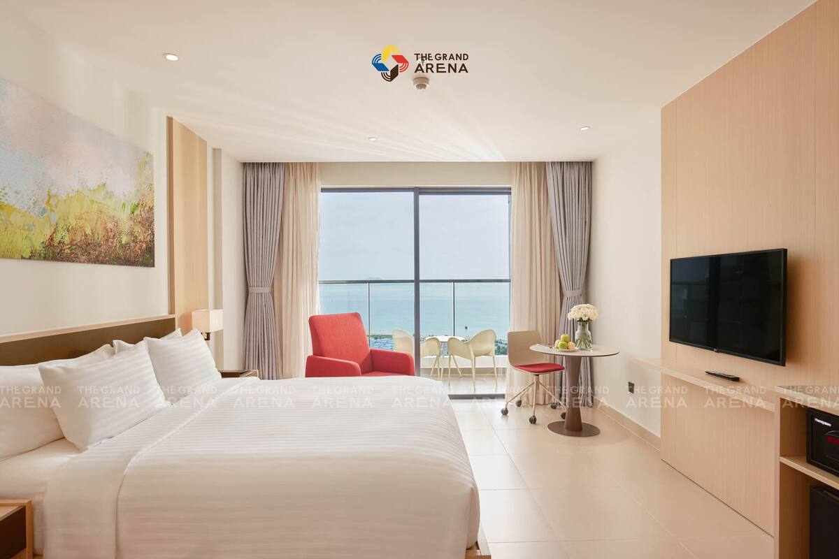 A hotel room with a large bed, a TV, a balcony with a view of the sea, and a red chair.
