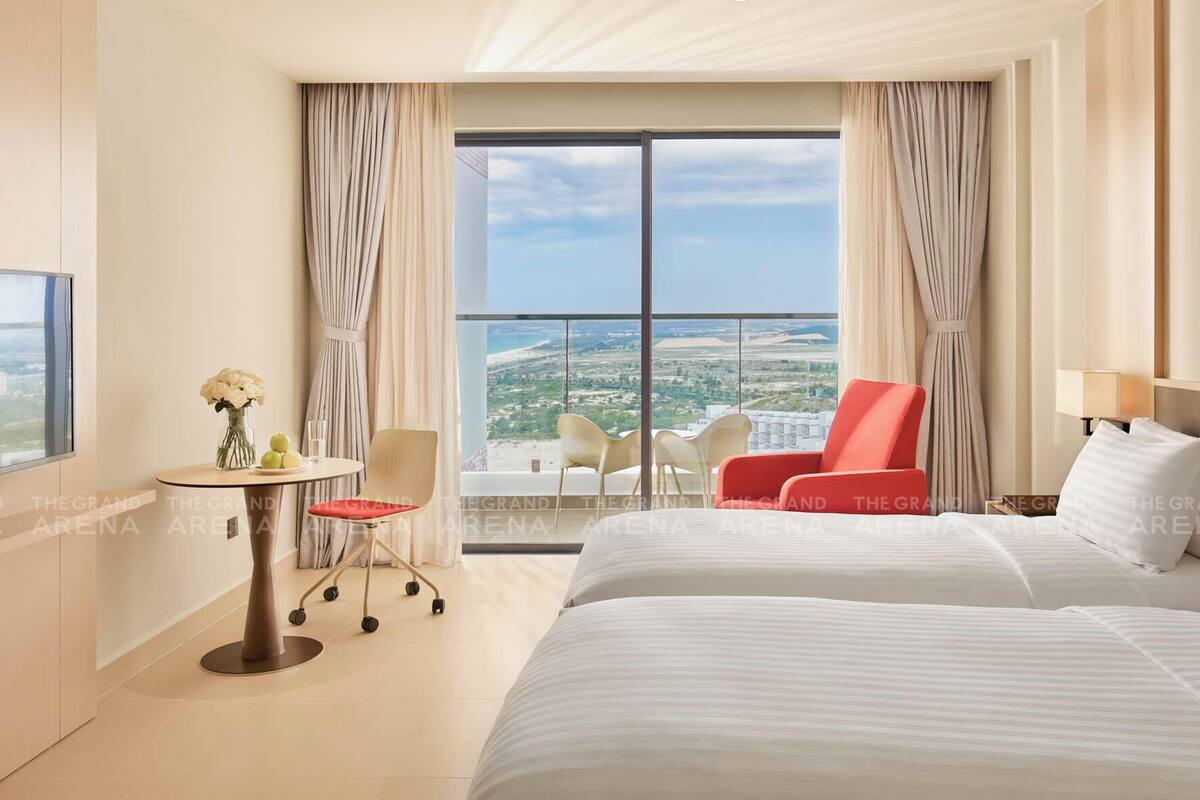A hotel room with a large bed, a desk with a chair, a small table with flowers, a red chair, and a view of the city through large windows.