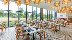 A spacious dining area with multiple tables and chairs, large windows offering a view of greenery, and hanging wicker-style pendant lights. - The Grand Arena Cam Ranh Hotel & Resort  (Bac Cam Ranh)