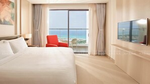 A hotel room with a large bed, a TV, a red chair, and a view of the ocean through the window. - The Grand Arena Cam Ranh Hotel & Resort  (Bac Cam Ranh)