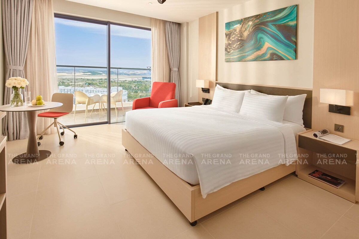 A hotel room with a large bed, a desk, a chair, a small table with flowers, a red chair, a painting, and a view of the city.
