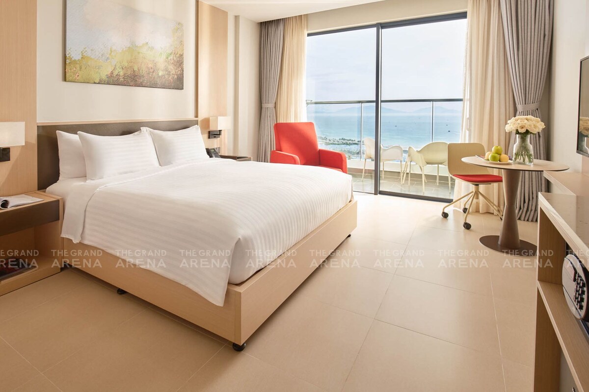 A hotel room with a large bed, a desk with a chair, a TV, and a view of the sea.
