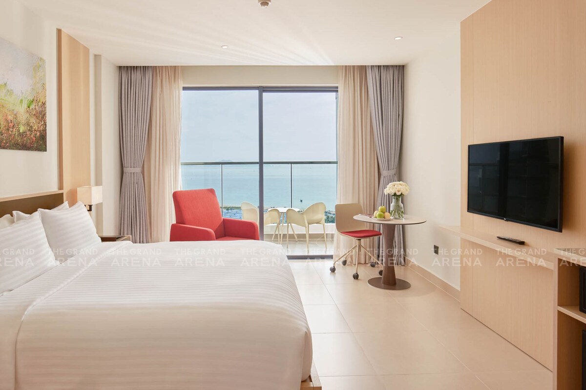 A hotel room with a large bed, a TV, a balcony with a view of the sea, and a red chair.