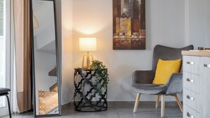 A modern room with a large mirror, a side table with a lamp and plant, a grey armchair with a yellow pillow, and a cityscape painting on the wall. - Acropolis Sandra Deluxe Apartment, Bill & John Apartments & Villas (Athens)