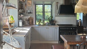 A kitchen with wooden beams, a hanging light fixture, a window with a view of greenery, and a dining area with a table and chairs.