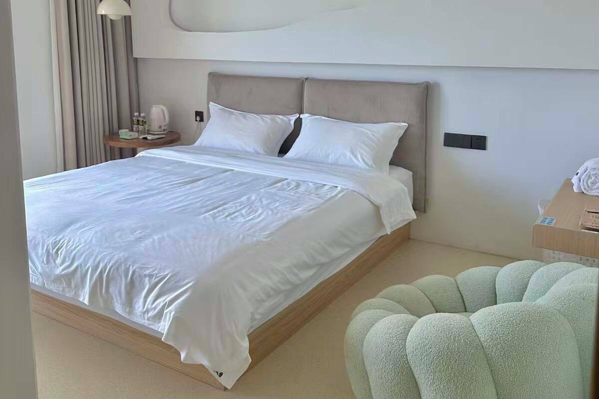 Una habitación de hotel moderna con una cama grande, una mesita de noche con lámpara y un otoman verde.