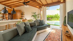 A modern living room with a large sectional sofa, a kitchen area with wooden cabinets, and a view of a swimming pool. - MALA Villa Balangan (Jimbaran)